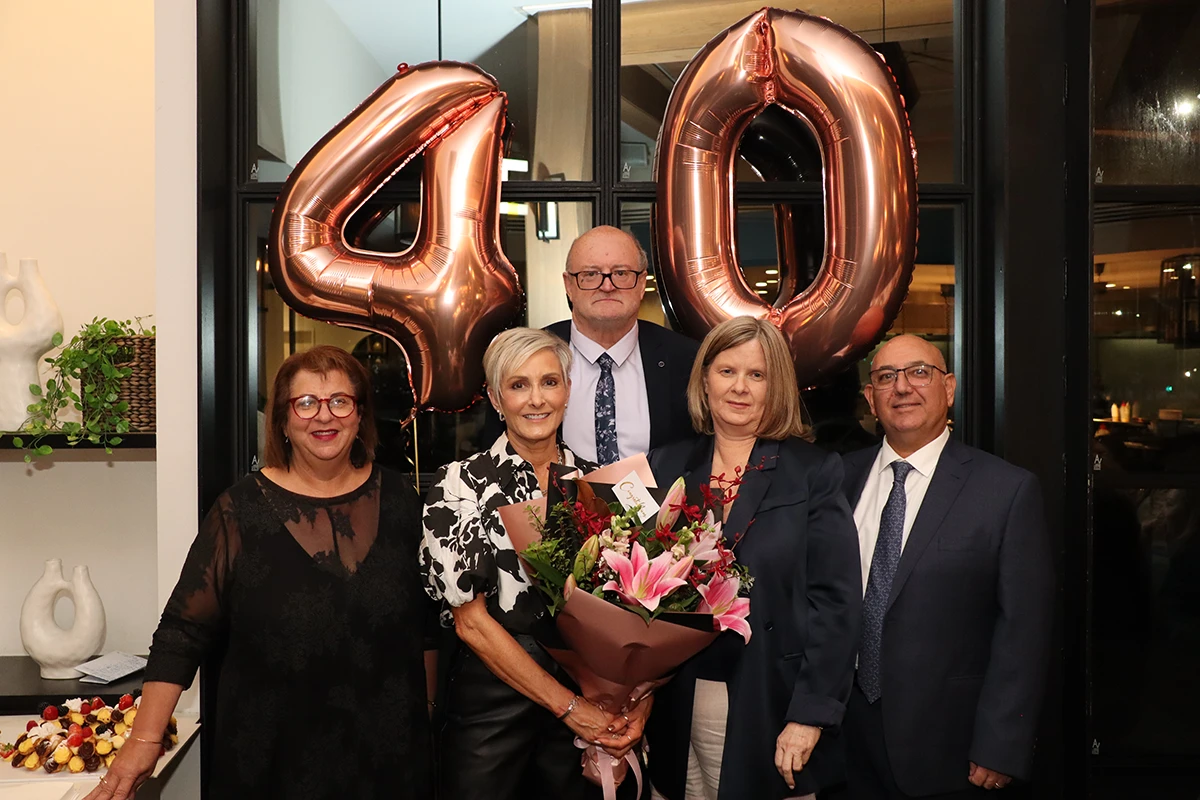 Five adults stand together indoors, posing for a photo in front of large rose-gold balloons shaped as the number 40. Christine Kennedy, at the center with a bouquet, is celebrated for 40 Years of Service. Everyone is dressed formally and smiling.